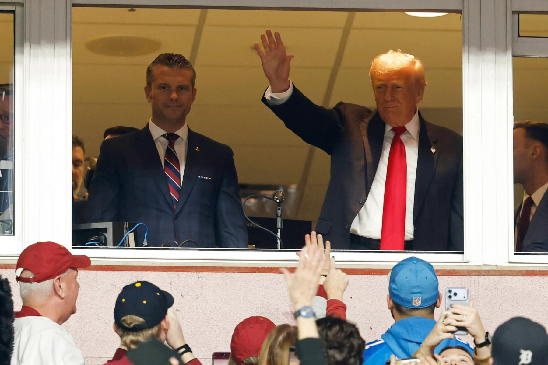 Zwei Männer stehen hinter einem Fenster über einer Menschenmenge. Der Mann rechts im dunklen Anzug und mit roter Krawatte, Donald Trump, winkt den Leuten unten zu. Einige im Vordergrund, vielleicht bei einem NFL-Spiel, schauen mit erhobenen Telefonen und Hüten in verschiedenen Farben auf. Diese Beschreibung wurde mit der FootballR KI automatisch generiert.