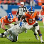 J.K. Dobbins - Ein Broncos-Spieler in Orange durchbricht einen Illegalen Tackle eines Verteidigers der Las Vegas Raiders, während seine Teamkollegen blocken. Der Spieler trägt den Football und läuft auf dem Spielfeld vorwärts, während im Hintergrund Schiedsrichter und andere Personen zu sehen sind. Diese Beschreibung wurde mit der FootballR KI automatisch generiert.