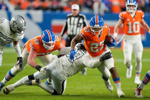 J.K. Dobbins - Ein Broncos-Spieler in Orange durchbricht einen Illegalen Tackle eines Verteidigers der Las Vegas Raiders, während seine Teamkollegen blocken. Der Spieler trägt den Football und läuft auf dem Spielfeld vorwärts, während im Hintergrund Schiedsrichter und andere Personen zu sehen sind. Diese Beschreibung wurde mit der FootballR KI automatisch generiert.