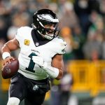 Jalen Hurts in einer weißen Eagles-Uniform mit der Nummer 1 läuft, während er den Ball hält. Er scheint konzentriert zu sein und blickt während eines NFC-Duells nach unten, wobei Fans und eine gelbe Absperrung im Stadionhintergrund verschwimmen. Diese Beschreibung wurde mit der FootballR KI automatisch generiert.