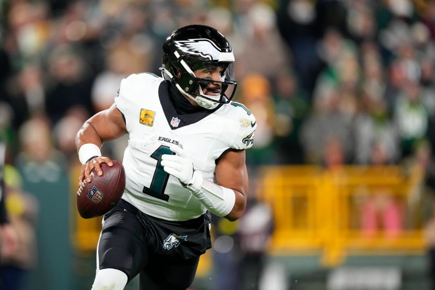 Jalen Hurts in einer weißen Eagles-Uniform mit der Nummer 1 läuft, während er den Ball hält. Er scheint konzentriert zu sein und blickt während eines NFC-Duells nach unten, wobei Fans und eine gelbe Absperrung im Stadionhintergrund verschwimmen. Diese Beschreibung wurde mit der FootballR KI automatisch generiert.