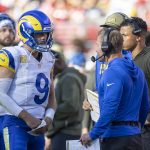 Ein Quarterback der Los Angeles Rams in voller Montur und mit Helm spricht mit Trainern in Blau und Braun an der Seitenlinie während eines Spiels der NFL Woche 12. Spieler und Mitarbeiter, von denen einige Kopfhörer tragen, stehen in der Nähe, während im Hintergrund eine unscharfe Menschenmenge zu sehen ist. Diese Beschreibung wurde mit der FootballR KI automatisch generiert.