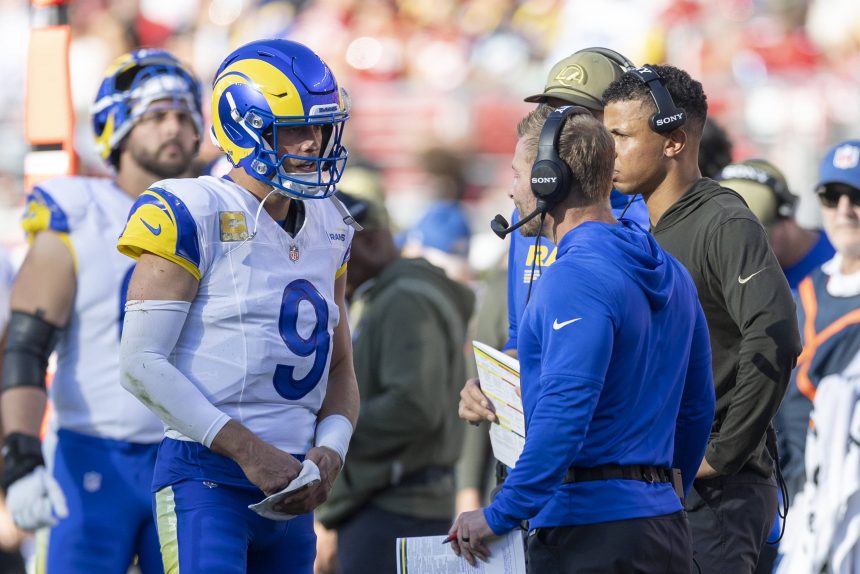 Ein Quarterback der Los Angeles Rams in voller Montur und mit Helm spricht mit Trainern in Blau und Braun an der Seitenlinie während eines Spiels der NFL Woche 12. Spieler und Mitarbeiter, von denen einige Kopfhörer tragen, stehen in der Nähe, während im Hintergrund eine unscharfe Menschenmenge zu sehen ist. Diese Beschreibung wurde mit der FootballR KI automatisch generiert.