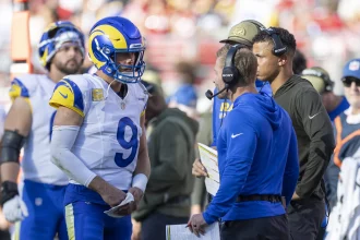 NFL Woche 4 - Ein Quarterback der Los Angeles Rams in voller Montur und mit Helm spricht mit Trainern in Blau und Braun an der Seitenlinie während eines Spiels der NFL Woche 12. Spieler und Mitarbeiter, von denen einige Kopfhörer tragen, stehen in der Nähe, während im Hintergrund eine unscharfe Menschenmenge zu sehen ist. Diese Beschreibung wurde mit der FootballR KI automatisch generiert.