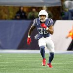 TreVeyon Henderson in einer marineblauen und silbernen Patriots-Uniform mit der Nummer 32 läuft auf einem grünen Feld. Er trägt rote Handschuhe, rote Stollenschuhe und einen silbernen Helm. Die unscharfe Seitenlinie deutet an, dass es um wichtige Touchdowns in einer Sportumgebung im Freien geht. Diese Beschreibung wurde mit der FootballR KI automatisch generiert.