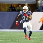TreVeyon Henderson in einer marineblauen und silbernen Patriots-Uniform mit der Nummer 32 läuft auf einem grünen Feld. Er trägt rote Handschuhe, rote Stollenschuhe und einen silbernen Helm. Die unscharfe Seitenlinie deutet an, dass es um wichtige Touchdowns in einer Sportumgebung im Freien geht. Diese Beschreibung wurde mit der FootballR KI automatisch generiert.