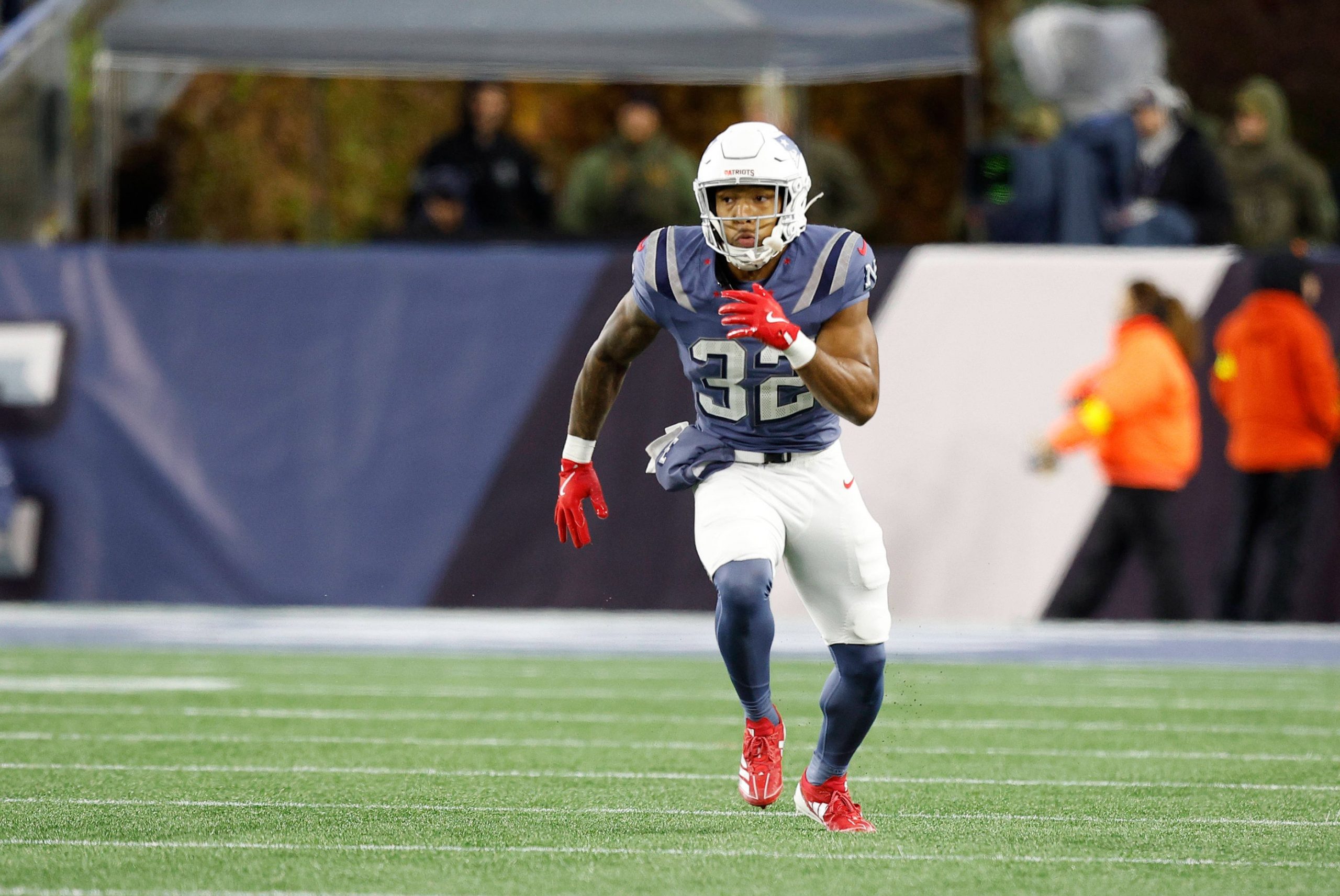 TreVeyon Henderson in einer marineblauen und silbernen Patriots-Uniform mit der Nummer 32 läuft auf einem grünen Feld. Er trägt rote Handschuhe, rote Stollenschuhe und einen silbernen Helm. Die unscharfe Seitenlinie deutet an, dass es um wichtige Touchdowns in einer Sportumgebung im Freien geht. Diese Beschreibung wurde mit der FootballR KI automatisch generiert.