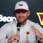 Matt Gay in weißem Sweatshirt und passender Baseballmütze mit kastanienbraunem und goldenem "W"-Logo, der möglicherweise über die NFL-Profis diskutiert oder ein Traum vom Madrid-Spiel erzählt, lächelt, während er bei einer Presseveranstaltung in ein Mikrofon spricht. Diese Beschreibung wurde mit der FootballR KI automatisch generiert.