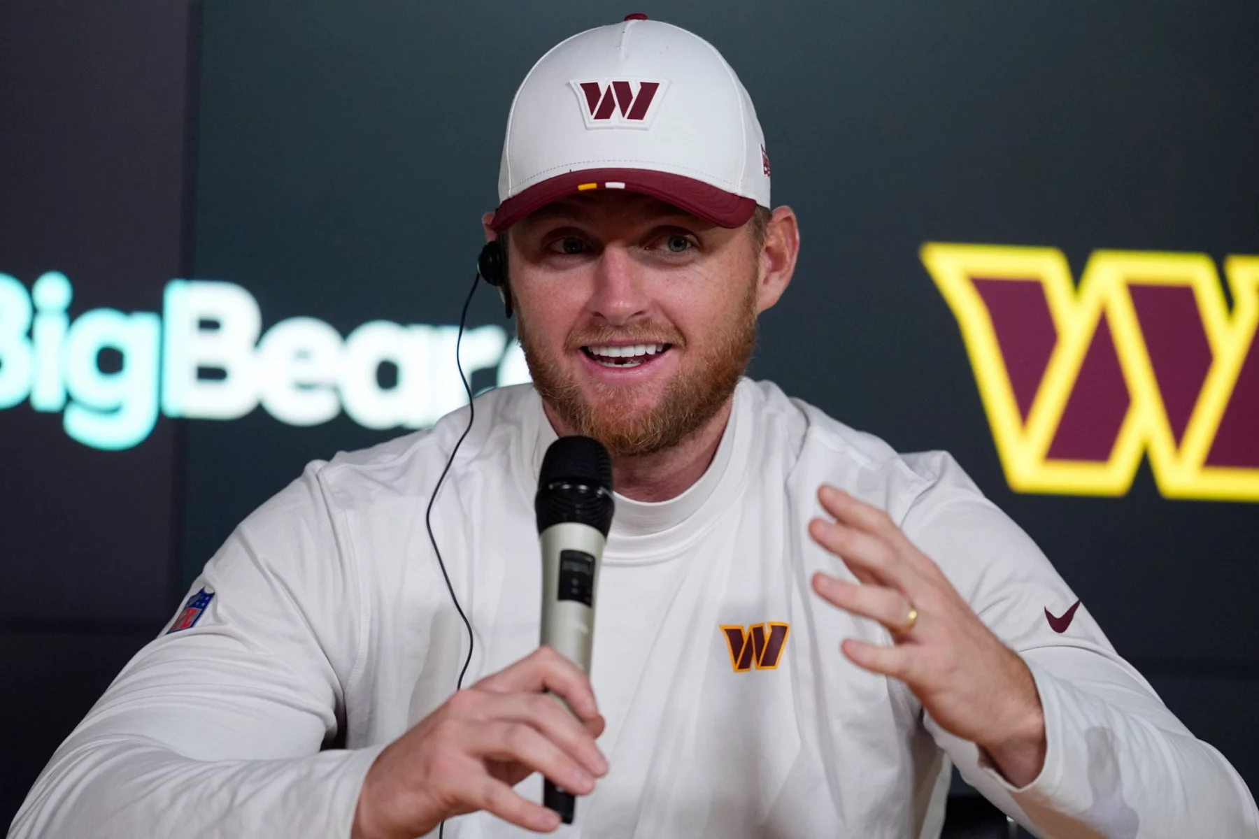 Matt Gay in weißem Sweatshirt und passender Baseballmütze mit kastanienbraunem und goldenem "W"-Logo, der möglicherweise über die NFL-Profis diskutiert oder ein Traum vom Madrid-Spiel erzählt, lächelt, während er bei einer Presseveranstaltung in ein Mikrofon spricht. Diese Beschreibung wurde mit der FootballR KI automatisch generiert.