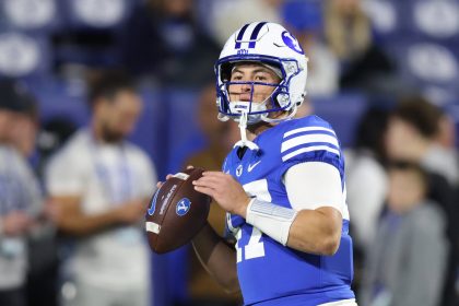 Ein Football-Spieler in einer blauen BYU-Uniform, weißem Helm und Armstulpen hält einen Football und bereitet sich auf den Wurf vor. Die unscharfen Zuschauer unterstreichen seine Konzentration bei diesem College-Football-Match und fangen die Energie von Woche 12 ein. Diese Beschreibung wurde mit der FootballR KI automatisch generiert.