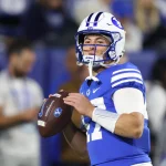 Ein Football-Spieler in einer blauen BYU-Uniform, weißem Helm und Armstulpen hält einen Football und bereitet sich auf den Wurf vor. Die unscharfen Zuschauer unterstreichen seine Konzentration bei diesem College-Football-Match und fangen die Energie von Woche 12 ein. Diese Beschreibung wurde mit der FootballR KI automatisch generiert.