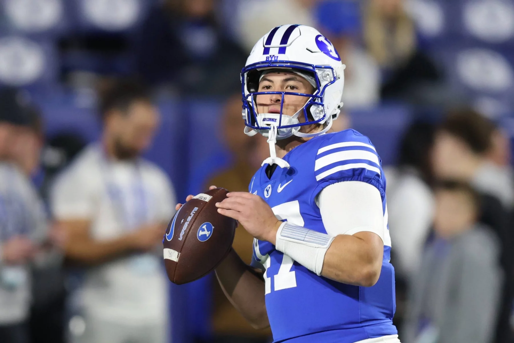 Ein Football-Spieler in einer blauen BYU-Uniform, weißem Helm und Armstulpen hält einen Football und bereitet sich auf den Wurf vor. Die unscharfen Zuschauer unterstreichen seine Konzentration bei diesem College-Football-Match und fangen die Energie von Woche 12 ein. Diese Beschreibung wurde mit der FootballR KI automatisch generiert.