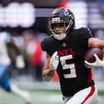 Drake London - Ein Footballspieler in einem schwarzen Trikot der Atlanta Falcons mit der Nummer 5 läuft mit dem Ball und schaut nach vorne. Er trägt einen Helm und Handschuhe, sein linker Arm hält den Football, während unscharfe Verteidiger in Weiß und Blau hinter ihm herlaufen - Drake London in Bewegung. Diese Beschreibung wurde mit der FootballR KI automatisch generiert.