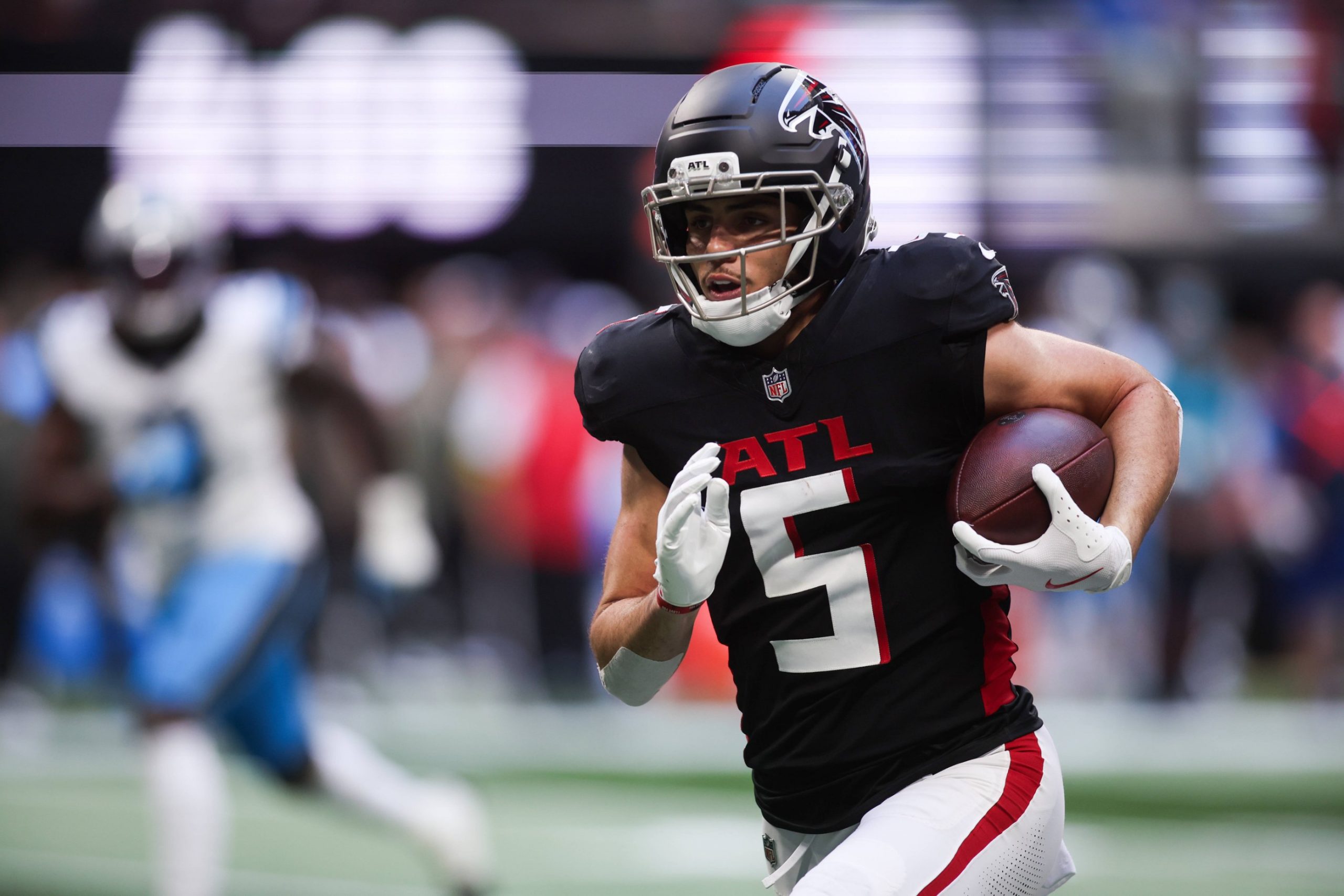 Drake London - Ein Footballspieler in einem schwarzen Trikot der Atlanta Falcons mit der Nummer 5 läuft mit dem Ball und schaut nach vorne. Er trägt einen Helm und Handschuhe, sein linker Arm hält den Football, während unscharfe Verteidiger in Weiß und Blau hinter ihm herlaufen - Drake London in Bewegung. Diese Beschreibung wurde mit der FootballR KI automatisch generiert.