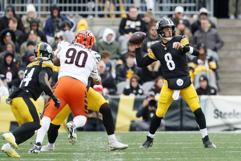 Woche 18 - Aaron Rodgers - Ein Steelers-Quarterback in einer schwarz-gelben Uniform (Nummer 8) bereitet sich darauf vor, den Ball zu werfen, während ein Verteidiger in einer weiß-orangefarbenen Uniform (Nummer 90) auf ihn zustürmt, was an klassische Aaron Rodgers-Momente erinnert. Fans und andere Spieler füllen den Hintergrund. Diese Beschreibung wurde mit der FootballR KI automatisch generiert.