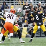 Aaron Rodgers - Ein Steelers-Quarterback in einer schwarz-gelben Uniform (Nummer 8) bereitet sich darauf vor, den Ball zu werfen, während ein Verteidiger in einer weiß-orangefarbenen Uniform (Nummer 90) auf ihn zustürmt, was an klassische Aaron Rodgers-Momente erinnert. Fans und andere Spieler füllen den Hintergrund. Diese Beschreibung wurde mit der FootballR KI automatisch generiert.