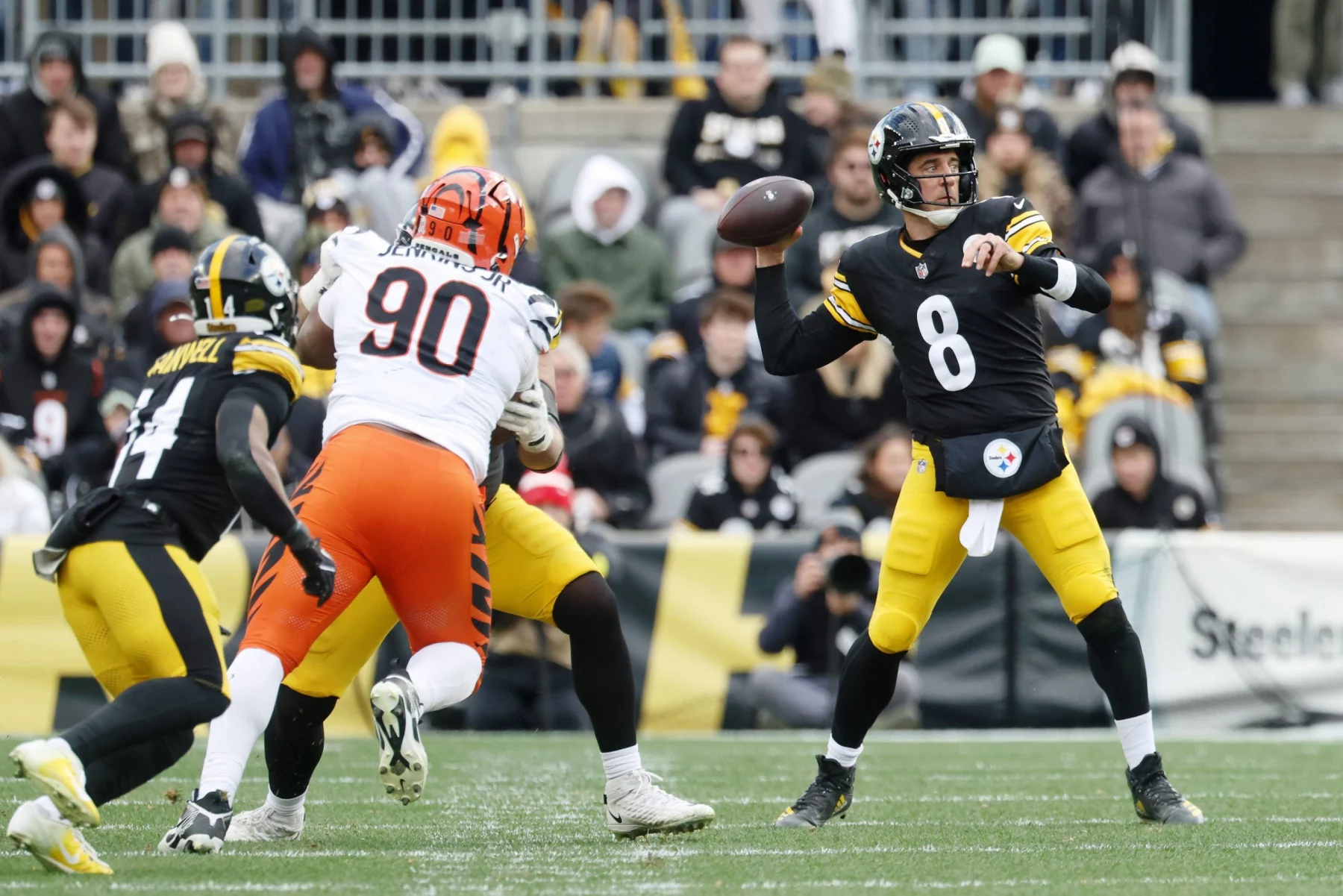 Woche 18 - Aaron Rodgers - Ein Steelers-Quarterback in einer schwarz-gelben Uniform (Nummer 8) bereitet sich darauf vor, den Ball zu werfen, während ein Verteidiger in einer weiß-orangefarbenen Uniform (Nummer 90) auf ihn zustürmt, was an klassische Aaron Rodgers-Momente erinnert. Fans und andere Spieler füllen den Hintergrund. Diese Beschreibung wurde mit der FootballR KI automatisch generiert.