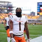 Ein Footballspieler der Cincinnati Bengals - möglicherweise Ja'Marr Chase - steht in einem weißen Trikot mit der Nummer 1, einer orangefarbenen Hose und einer schwarzen Skimaske auf dem Spielfeld. Die Sitze im Stadion dahinter sind überwiegend gelb, der Himmel ist bedeckt. Diese Beschreibung wurde mit der FootballR KI automatisch generiert.