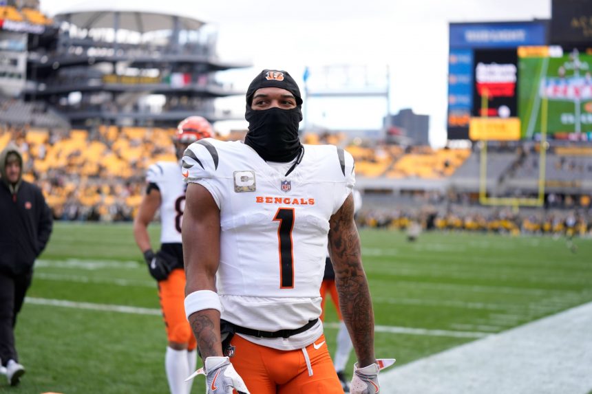Ein Footballspieler der Cincinnati Bengals - möglicherweise Ja'Marr Chase - steht in einem weißen Trikot mit der Nummer 1, einer orangefarbenen Hose und einer schwarzen Skimaske auf dem Spielfeld. Die Sitze im Stadion dahinter sind überwiegend gelb, der Himmel ist bedeckt. Diese Beschreibung wurde mit der FootballR KI automatisch generiert.