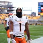 Ein Footballspieler der Cincinnati Bengals - möglicherweise Ja'Marr Chase - steht in einem weißen Trikot mit der Nummer 1, einer orangefarbenen Hose und einer schwarzen Skimaske auf dem Spielfeld. Die Sitze im Stadion dahinter sind überwiegend gelb, der Himmel ist bedeckt. Diese Beschreibung wurde mit der FootballR KI automatisch generiert.