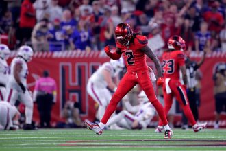 Ein Footballspieler in einem roten Texans-Trikot mit der Nummer 2 ballt die Faust und blickt jubelnd auf das Spielfeld. Im Hintergrund sind andere Spieler und eine jubelnde Menge zu sehen, die Stadionbeleuchtung erhellt die Szene. Diese Beschreibung wurde mit der FootballR KI automatisch generiert.