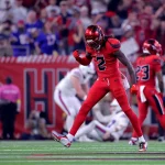 Ein Footballspieler in einem roten Texans-Trikot mit der Nummer 2 ballt die Faust und blickt jubelnd auf das Spielfeld. Im Hintergrund sind andere Spieler und eine jubelnde Menge zu sehen, die Stadionbeleuchtung erhellt die Szene. Diese Beschreibung wurde mit der FootballR KI automatisch generiert.