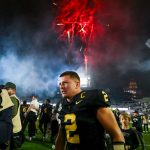 Diego Pavia - Ein Footballspieler in einem schwarz-goldenen Trikot mit der Nummer 2 feiert nachts auf dem Spielfeld während der College Week 13, umgeben von Fotografen und Fans. Rote Feuerwerkskörper explodieren in den rauchigen Himmel darüber und tragen zur festlichen Atmosphäre bei. Diese Beschreibung wurde mit der FootballR KI automatisch generiert.