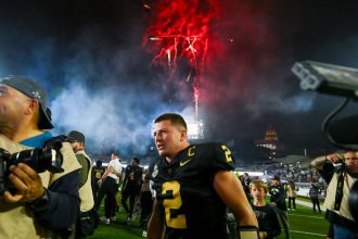 Diego Pavia - Ein Footballspieler in einem schwarz-goldenen Trikot mit der Nummer 2 feiert nachts auf dem Spielfeld während der College Week 13, umgeben von Fotografen und Fans. Rote Feuerwerkskörper explodieren in den rauchigen Himmel darüber und tragen zur festlichen Atmosphäre bei. Diese Beschreibung wurde mit der FootballR KI automatisch generiert.