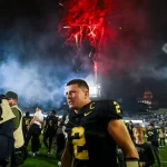 Diego Pavia - Ein Footballspieler in einem schwarz-goldenen Trikot mit der Nummer 2 feiert nachts auf dem Spielfeld während der College Week 13, umgeben von Fotografen und Fans. Rote Feuerwerkskörper explodieren in den rauchigen Himmel darüber und tragen zur festlichen Atmosphäre bei. Diese Beschreibung wurde mit der FootballR KI automatisch generiert.