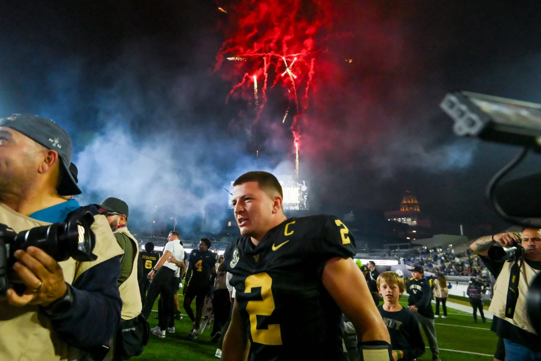Diego Pavia - Ein Footballspieler in einem schwarz-goldenen Trikot mit der Nummer 2 feiert nachts auf dem Spielfeld während der College Week 13, umgeben von Fotografen und Fans. Rote Feuerwerkskörper explodieren in den rauchigen Himmel darüber und tragen zur festlichen Atmosphäre bei. Diese Beschreibung wurde mit der FootballR KI automatisch generiert.