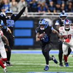 Jahmyr Gibbs - Woche 12 - Ein Footballspieler in einem schwarzen Trikot der Detroit Lions läuft mit dem Ball, verfolgt von mehreren Giants-Verteidigern in Weiß. Mitspieler und Gegner laufen über das grüne Feld, während die Fans im Hintergrund von der Tribüne aus zusehen. Diese Beschreibung wurde mit der FootballR KI automatisch generiert.