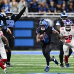 Jahmyr Gibbs - Woche 12 - Ein Footballspieler in einem schwarzen Trikot der Detroit Lions läuft mit dem Ball, verfolgt von mehreren Giants-Verteidigern in Weiß. Mitspieler und Gegner laufen über das grüne Feld, während die Fans im Hintergrund von der Tribüne aus zusehen. Diese Beschreibung wurde mit der FootballR KI automatisch generiert.