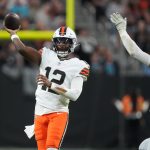 Ein Footballspieler, Shedeur Sanders, trägt ein weißes Trikot mit der Nummer 12 und orangefarbenen Streifen und bereitet sich darauf vor, einen Pass zu werfen. Er befindet sich mitten im Wurf und sieht sich einem Verteidiger in Schwarz gegenüber, der sich auf dem Spielfeld ausstreckt, während das Publikum im Hintergrund verschwommen ist. Diese Beschreibung wurde mit der FootballR KI automatisch generiert.