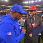 Auf dem Football-Feld schüttelt ein Trainer in blauer NFL-Jacke und Mütze dem Bucs-Quarterback Baker Mayfield die Hand. Er trägt eine rote Mütze, einen grauen Buccaneers-Kapuzenpulli und seinen Arm in einer Schlinge - ein Moment, in dem die Zuschauer und Fotografen unter den Stadionlichtern auf den neuesten Stand der Verletzungen gebracht werden. Diese Beschreibung wurde mit der FootballR KI automatisch generiert.