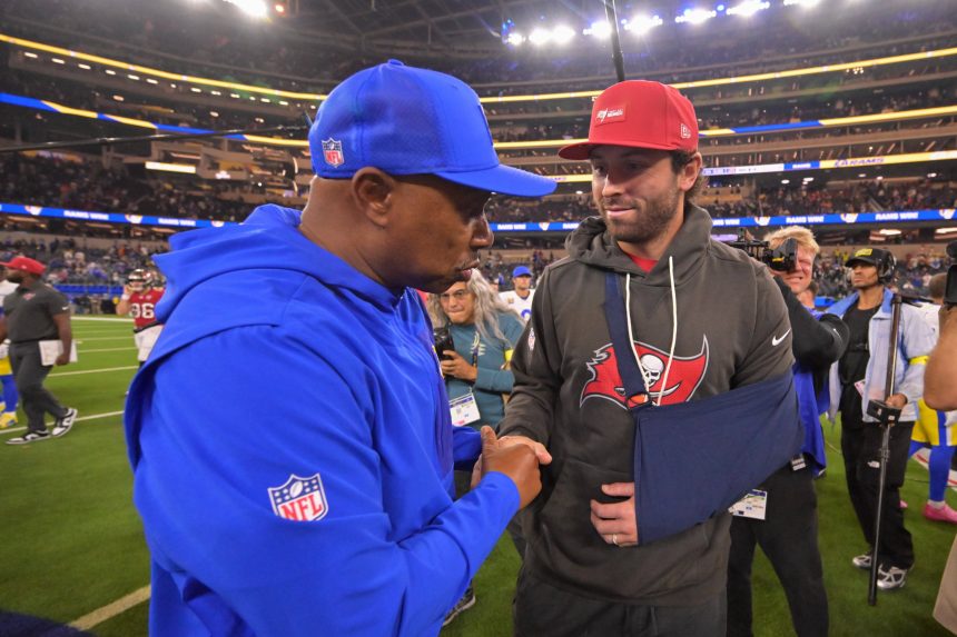 Auf dem Football-Feld schüttelt ein Trainer in blauer NFL-Jacke und Mütze dem Bucs-Quarterback Baker Mayfield die Hand. Er trägt eine rote Mütze, einen grauen Buccaneers-Kapuzenpulli und seinen Arm in einer Schlinge - ein Moment, in dem die Zuschauer und Fotografen unter den Stadionlichtern auf den neuesten Stand der Verletzungen gebracht werden. Diese Beschreibung wurde mit der FootballR KI automatisch generiert.