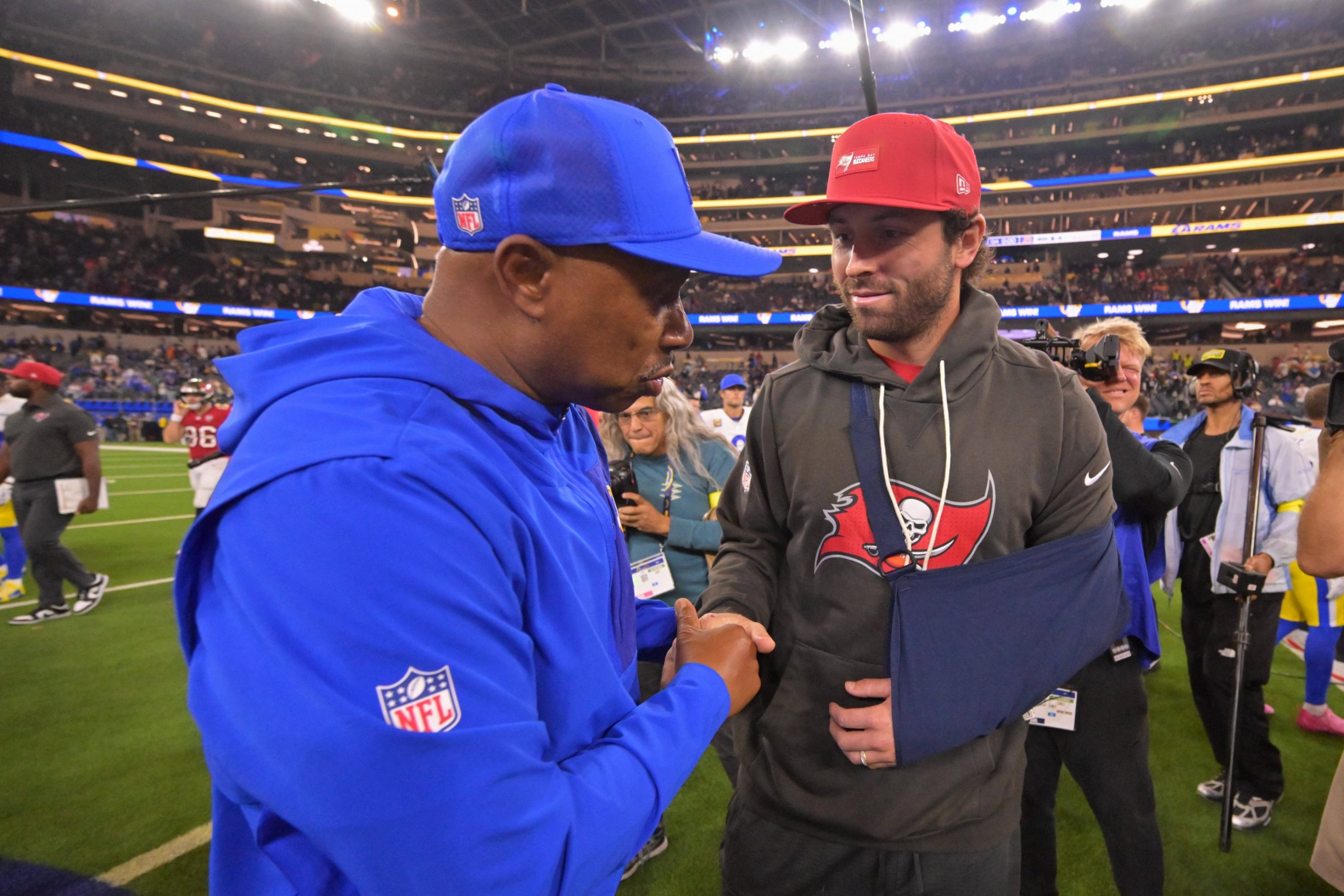 Auf dem Football-Feld schüttelt ein Trainer in blauer NFL-Jacke und Mütze dem Bucs-Quarterback Baker Mayfield die Hand. Er trägt eine rote Mütze, einen grauen Buccaneers-Kapuzenpulli und seinen Arm in einer Schlinge - ein Moment, in dem die Zuschauer und Fotografen unter den Stadionlichtern auf den neuesten Stand der Verletzungen gebracht werden. Diese Beschreibung wurde mit der FootballR KI automatisch generiert.