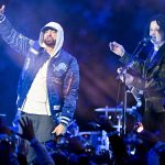 Zwei Musiker treten bei Nacht auf der Bühne auf. Der Künstler auf der linken Seite, in einer blauen Detroit Lions-Jacke und einem weißen Kapuzenpulli, verkörpert den Stolz auf Detroit wie Eminem, während der Musiker auf der rechten Seite singt und Gitarre spielt, während die Menge im Vordergrund die Hände hebt. Diese Beschreibung wurde mit der FootballR KI automatisch generiert.