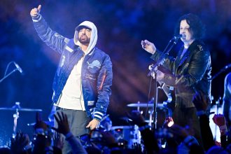 Zwei Musiker treten bei Nacht auf der Bühne auf. Der Künstler auf der linken Seite, in einer blauen Detroit Lions-Jacke und einem weißen Kapuzenpulli, verkörpert den Stolz auf Detroit wie Eminem, während der Musiker auf der rechten Seite singt und Gitarre spielt, während die Menge im Vordergrund die Hände hebt. Diese Beschreibung wurde mit der FootballR KI automatisch generiert.