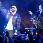 Zwei Musiker treten bei Nacht auf der Bühne auf. Der Künstler auf der linken Seite, in einer blauen Detroit Lions-Jacke und einem weißen Kapuzenpulli, verkörpert den Stolz auf Detroit wie Eminem, während der Musiker auf der rechten Seite singt und Gitarre spielt, während die Menge im Vordergrund die Hände hebt. Diese Beschreibung wurde mit der FootballR KI automatisch generiert.