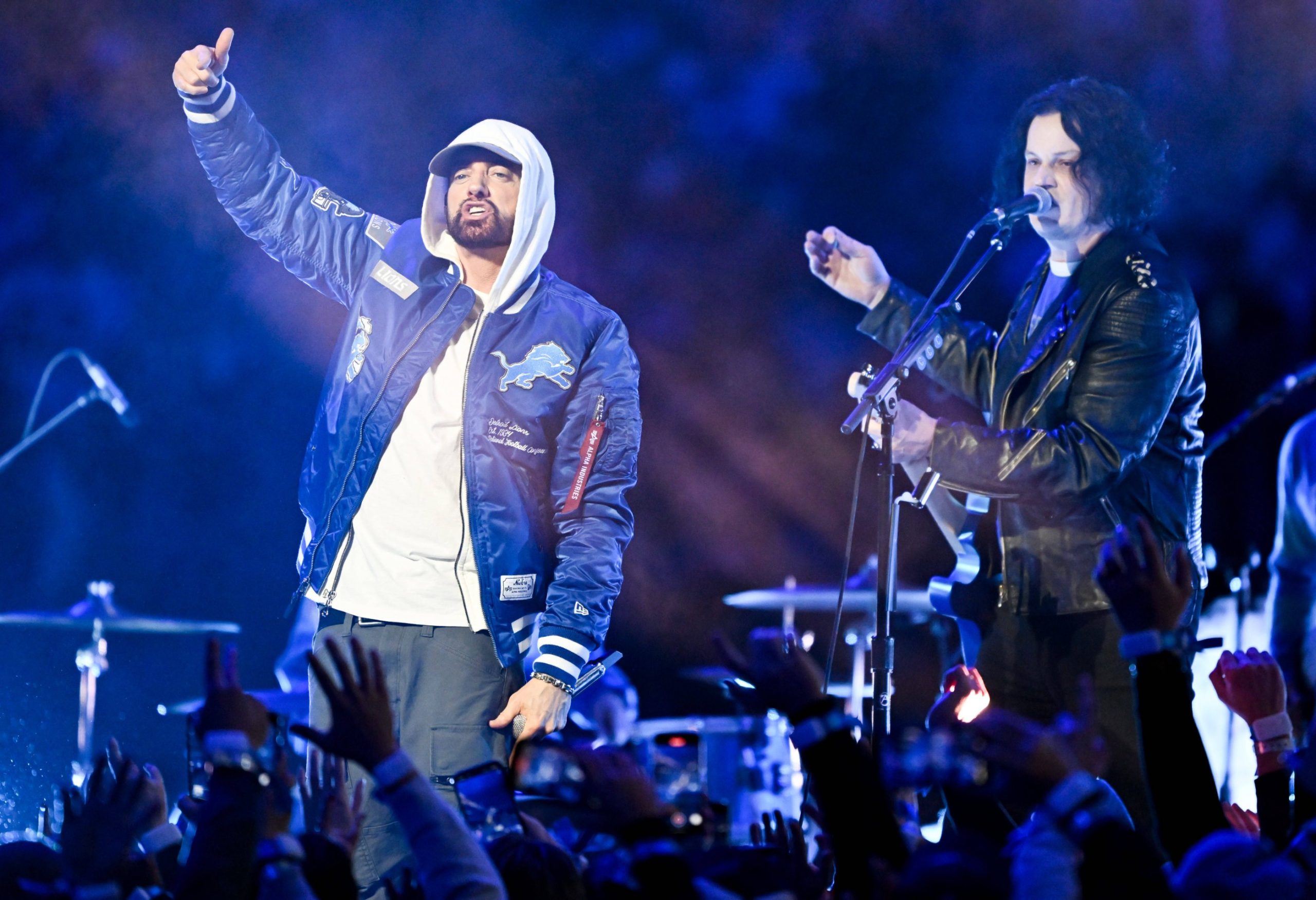 Zwei Musiker treten bei Nacht auf der Bühne auf. Der Künstler auf der linken Seite, in einer blauen Detroit Lions-Jacke und einem weißen Kapuzenpulli, verkörpert den Stolz auf Detroit wie Eminem, während der Musiker auf der rechten Seite singt und Gitarre spielt, während die Menge im Vordergrund die Hände hebt. Diese Beschreibung wurde mit der FootballR KI automatisch generiert.