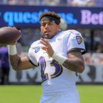 Jaire Alexander - Ein Footballspieler in einem weißen Baltimore Ravens-Trikot mit der Nummer 23 hält einen Football und bereitet sich auf den Wurf vor. Er trägt ein schwarzes Stirnband und steht auf einem Feld, mit unscharfen Philadelphia Eagles-Schildern und Zuschauern im Hintergrund. Diese Beschreibung wurde mit der FootballR KI automatisch generiert.