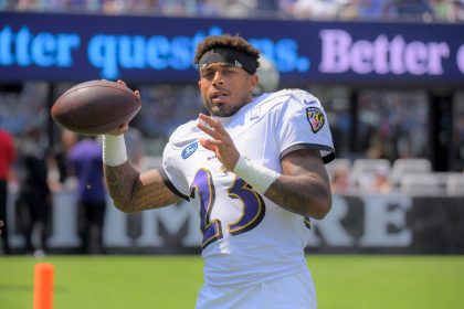 Jaire Alexander - Ein Footballspieler in einem weißen Baltimore Ravens-Trikot mit der Nummer 23 hält einen Football und bereitet sich auf den Wurf vor. Er trägt ein schwarzes Stirnband und steht auf einem Feld, mit unscharfen Philadelphia Eagles-Schildern und Zuschauern im Hintergrund. Diese Beschreibung wurde mit der FootballR KI automatisch generiert.