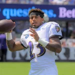 Jaire Alexander - Ein Footballspieler in einem weißen Baltimore Ravens-Trikot mit der Nummer 23 hält einen Football und bereitet sich auf den Wurf vor. Er trägt ein schwarzes Stirnband und steht auf einem Feld, mit unscharfen Philadelphia Eagles-Schildern und Zuschauern im Hintergrund. Diese Beschreibung wurde mit der FootballR KI automatisch generiert.
