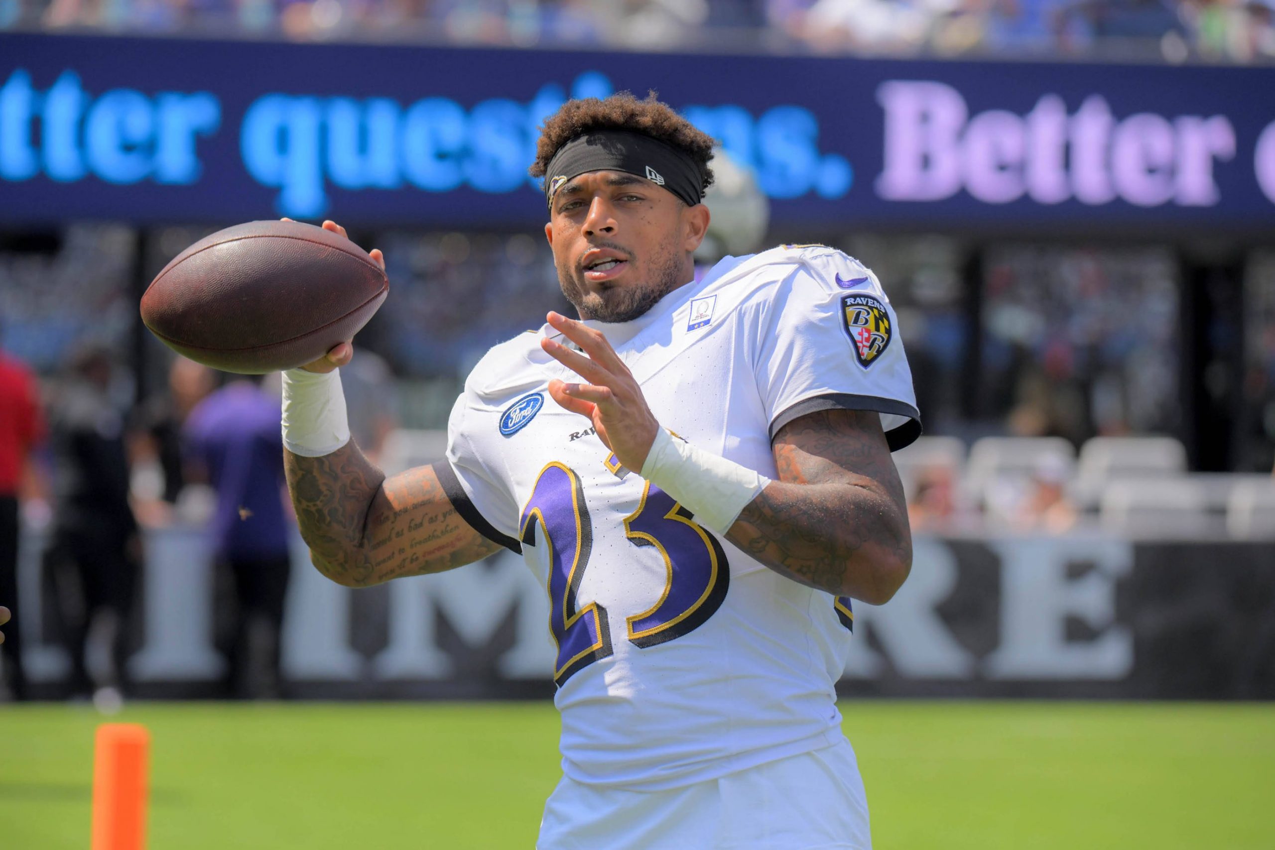 Jaire Alexander - Ein Footballspieler in einem weißen Baltimore Ravens-Trikot mit der Nummer 23 hält einen Football und bereitet sich auf den Wurf vor. Er trägt ein schwarzes Stirnband und steht auf einem Feld, mit unscharfen Philadelphia Eagles-Schildern und Zuschauern im Hintergrund. Diese Beschreibung wurde mit der FootballR KI automatisch generiert.