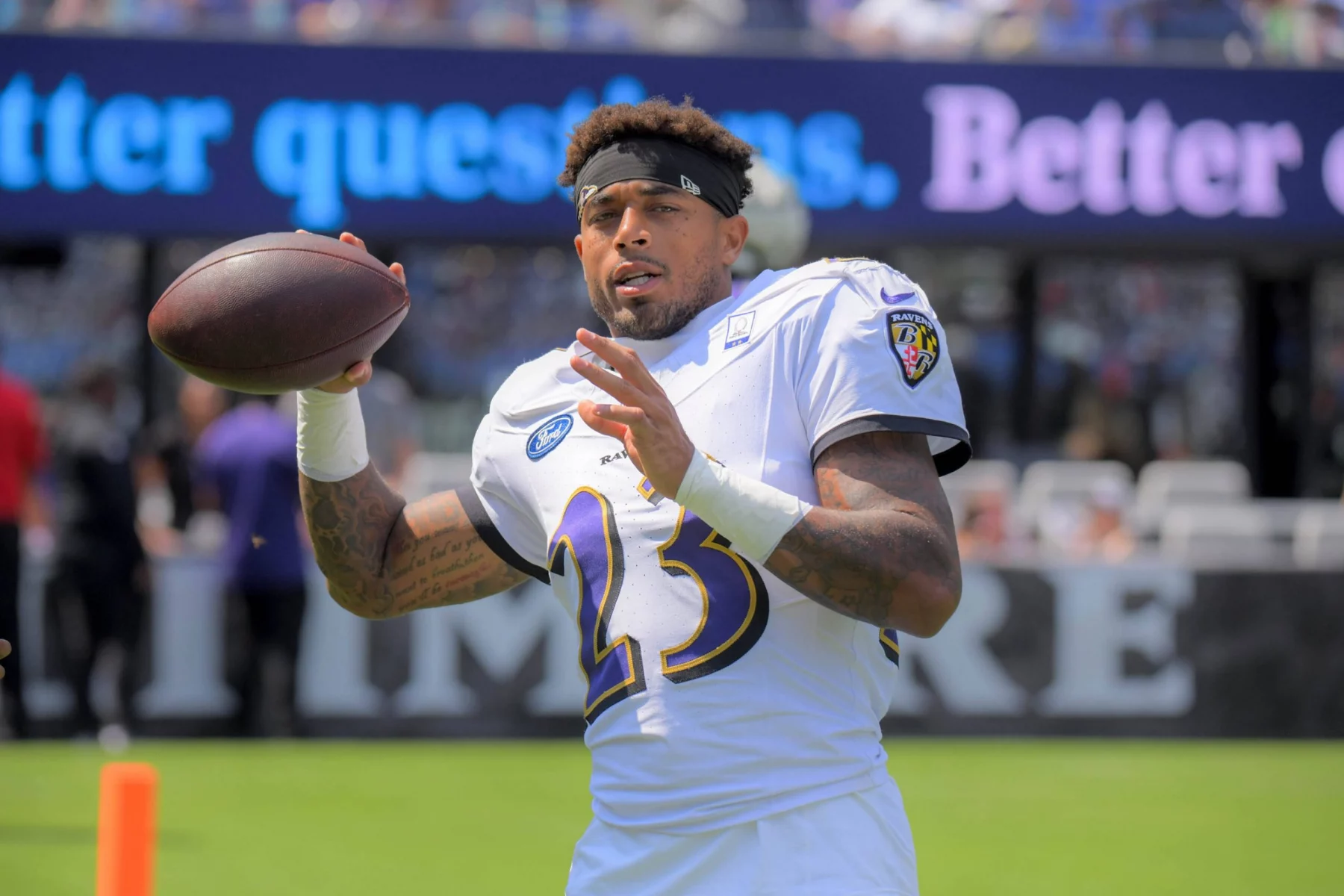 Jaire Alexander - Ein Footballspieler in einem weißen Baltimore Ravens-Trikot mit der Nummer 23 hält einen Football und bereitet sich auf den Wurf vor. Er trägt ein schwarzes Stirnband und steht auf einem Feld, mit unscharfen Philadelphia Eagles-Schildern und Zuschauern im Hintergrund. Diese Beschreibung wurde mit der FootballR KI automatisch generiert.