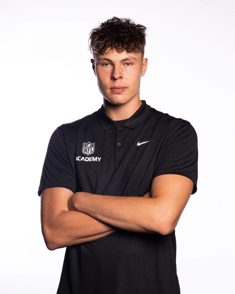 Bruno Werner - Ein junger Mann mit lockigem braunem Haar, der ein schwarzes Nike-Poloshirt mit "NFL Academy"-Logo trägt, steht selbstbewusst mit verschränkten Armen da und verkörpert den Ehrgeiz der NFL Academy Absolventen, die auf US-Colleges abzielen. Der Hintergrund ist schlicht weiß, die Beleuchtung hell und gleichmäßig. Diese Beschreibung wurde mit der FootballR KI automatisch generiert.