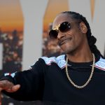Ein Mann mit langen Zöpfen, der eine schwarze Sonnenbrille, eine goldene Kette und einen schwarzen Pullover mit weißen und blauen Mustern trägt, lächelt und gestikuliert mit einer Hand. Die Stimmung erinnert an Snoop Dogg bei einer NFL-Weihnachtshalbzeitshow vor dem Hintergrund einer untergehenden Stadtkulisse. Diese Beschreibung wurde mit der FootballR KI automatisch generiert.