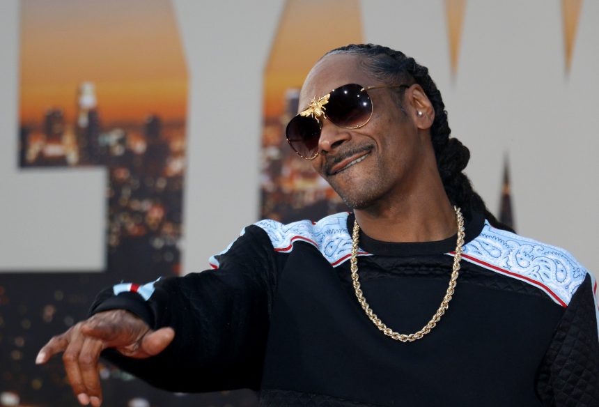 Ein Mann mit langen Zöpfen, der eine schwarze Sonnenbrille, eine goldene Kette und einen schwarzen Pullover mit weißen und blauen Mustern trägt, lächelt und gestikuliert mit einer Hand. Die Stimmung erinnert an Snoop Dogg bei einer NFL-Weihnachtshalbzeitshow vor dem Hintergrund einer untergehenden Stadtkulisse. Diese Beschreibung wurde mit der FootballR KI automatisch generiert.