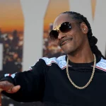 Ein Mann mit langen Zöpfen, der eine schwarze Sonnenbrille, eine goldene Kette und einen schwarzen Pullover mit weißen und blauen Mustern trägt, lächelt und gestikuliert mit einer Hand. Die Stimmung erinnert an Snoop Dogg bei einer NFL-Weihnachtshalbzeitshow vor dem Hintergrund einer untergehenden Stadtkulisse. Diese Beschreibung wurde mit der FootballR KI automatisch generiert.