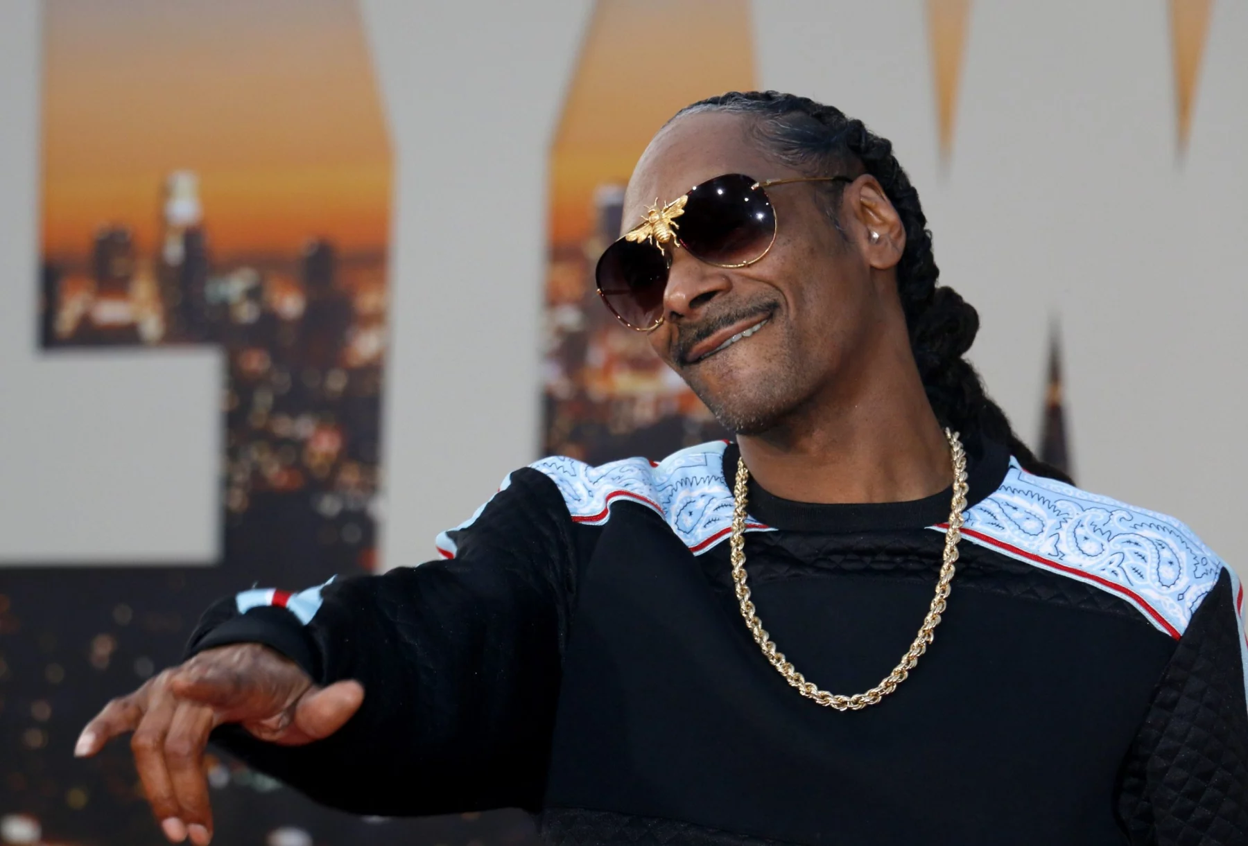 Ein Mann mit langen Zöpfen, der eine schwarze Sonnenbrille, eine goldene Kette und einen schwarzen Pullover mit weißen und blauen Mustern trägt, lächelt und gestikuliert mit einer Hand. Die Stimmung erinnert an Snoop Dogg bei einer NFL-Weihnachtshalbzeitshow vor dem Hintergrund einer untergehenden Stadtkulisse. Diese Beschreibung wurde mit der FootballR KI automatisch generiert.